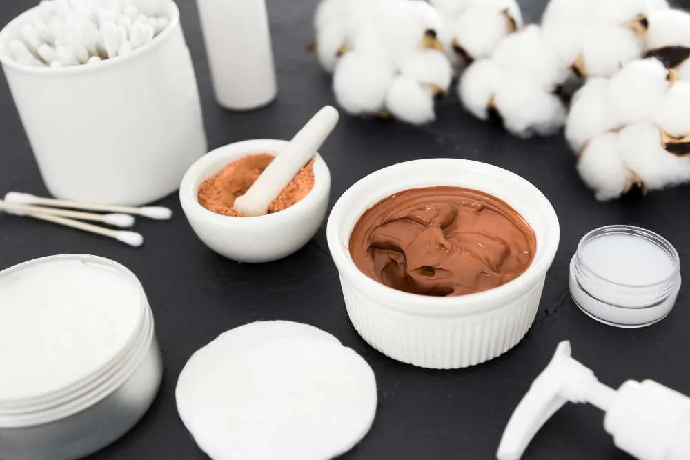 Clay masks, cotton and skincare pots laid out on a dark surface.