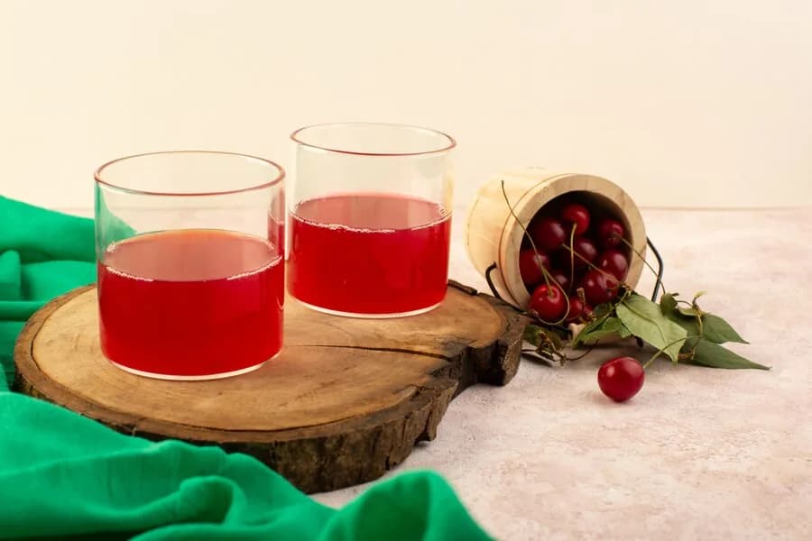 Two glasses of red fruit drink beside a cup of cherries on a wooden board.