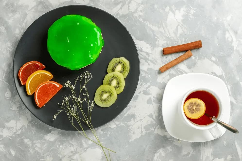 A green jelly dessert with orange slices and kiwi, beside a cup of lemon tea.