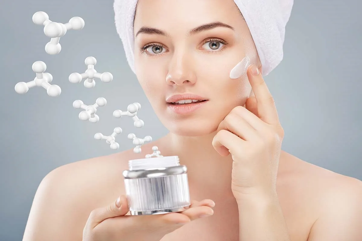 A woman with a towel wrap touching cream to her cheek while holding the jar.