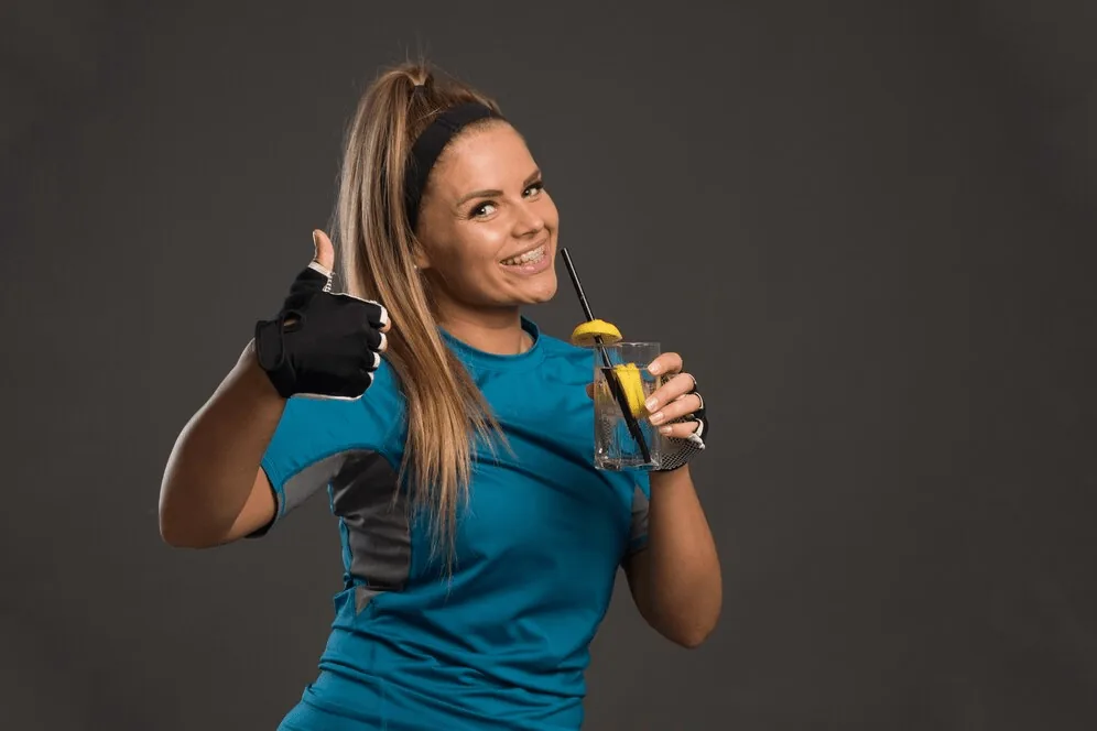 A smiling woman in sportswear holding a nutrition drink and giving a thumbs-up.