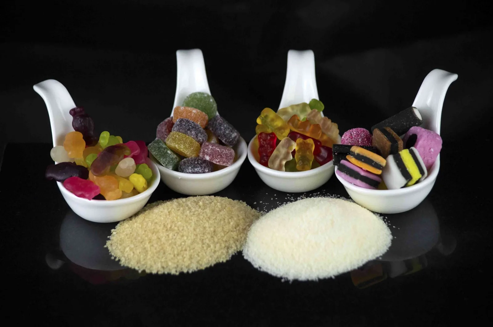 Gummy sweets in four porcelain spoons above two piles of gelatin granules and powder.