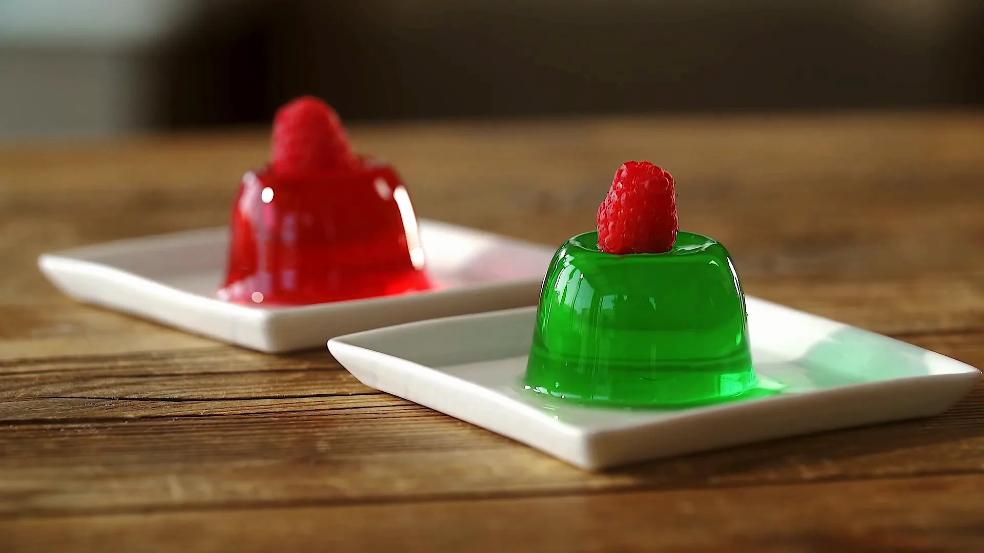 Red and green gelatin desserts, each topped with a raspberry, on white plates.