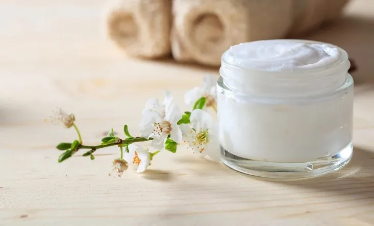An open jar of white cream beside sprigs of blossom.