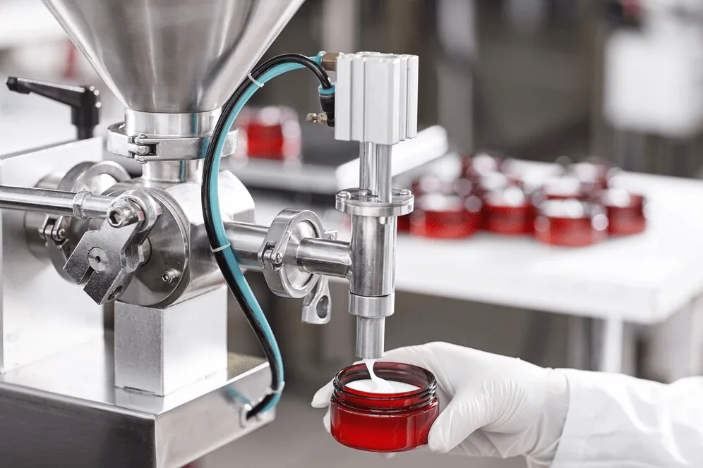 A filling machine dispensing red cream into jars on a cosmetics production line.