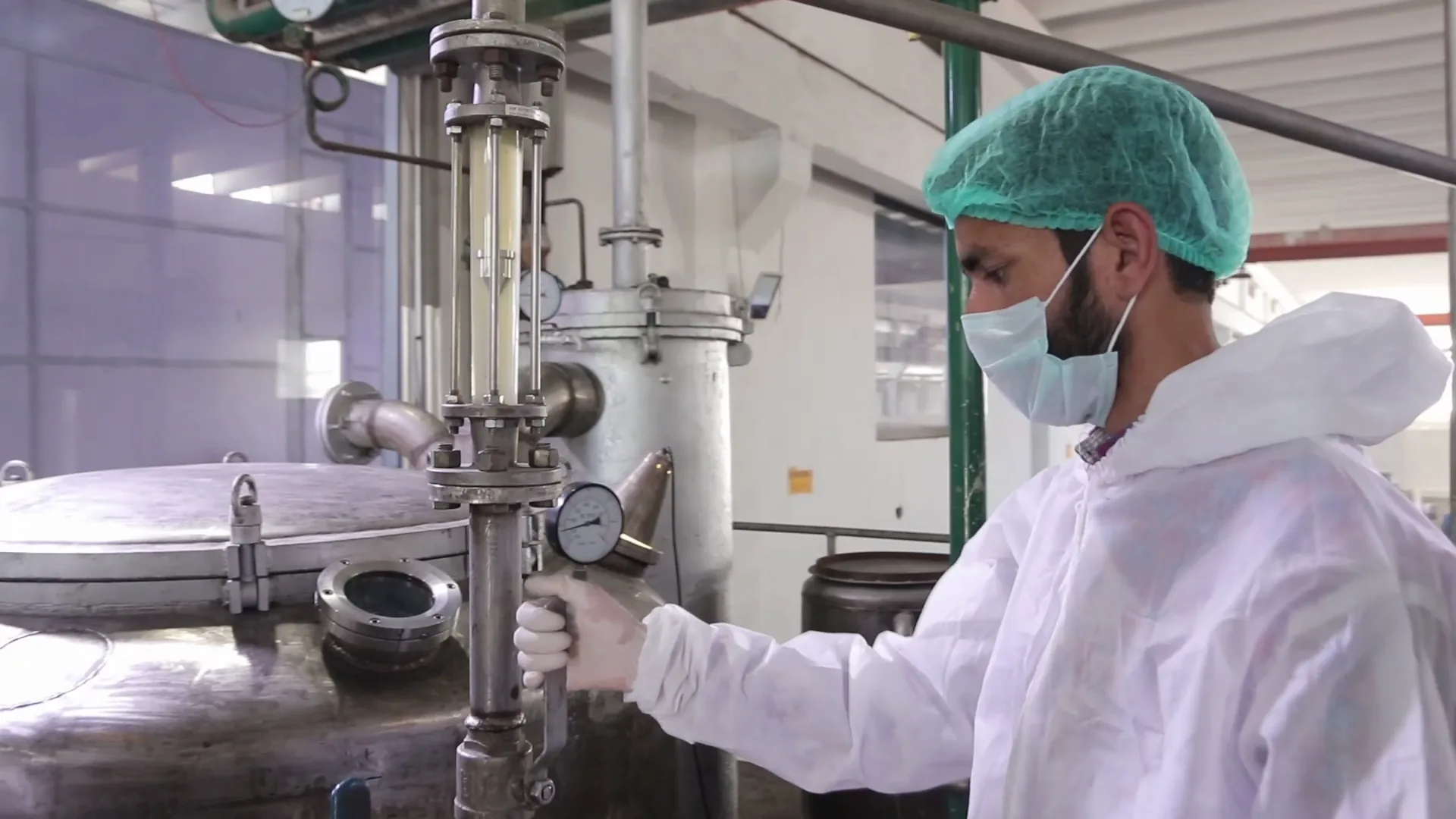 A Sadiq Gelatin operator in hairnet, mask and cleanroom suit adjusting a valve on a stainless-steel reactor.