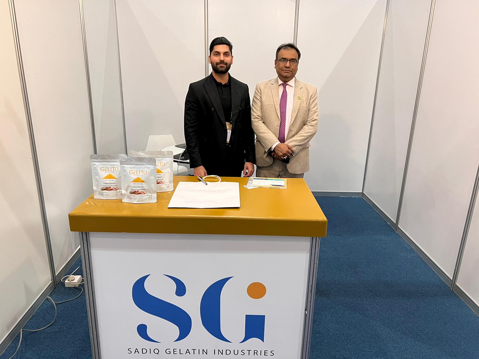 Two members of the Sadiq Gelatin team behind the stand counter with retail packs of Halal gelatin.